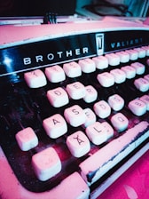 A vintage typewriter with the brand name 'Brother Valiant' is displayed at an angle. The keys are white with black lettering and numbers, showing signs of wear and age with a slightly dusty appearance. The typewriter's body has a retro aesthetic, emphasizing a sense of nostalgia.