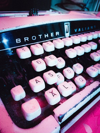 A vintage typewriter with the brand name 'Brother Valiant' is displayed at an angle. The keys are white with black lettering and numbers, showing signs of wear and age with a slightly dusty appearance. The typewriter's body has a retro aesthetic, emphasizing a sense of nostalgia.