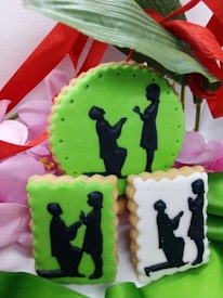 Three cookies displaying silhouettes of a couple, where one figure is kneeling and proposing to the other. The cookies are decorated with green and white icing, with floral and ribbon adornments in the background.