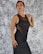 A woman with red hair poses confidently wearing a black dress that has mesh detailing. She stands against a textured black and white backdrop, showcasing her attire with one hand on her hip and the other gently placed near her neck.