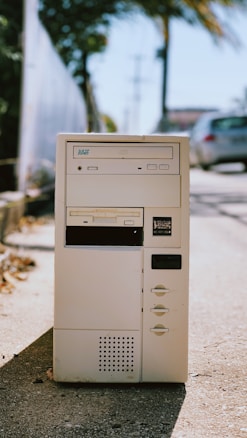 A vintage computer tower with a beige color, featuring a floppy disk drive, CD drive, and multiple buttons, placed outdoors on a sunlit sidewalk. The background includes blurred elements of greenery and parked cars.