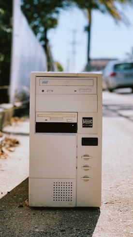A vintage computer tower with a beige color, featuring a floppy disk drive, CD drive, and multiple buttons, placed outdoors on a sunlit sidewalk. The background includes blurred elements of greenery and parked cars.