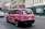 A pink taxi with environmental advertising is driving on a city street. The vehicle has the words 'This cab is green' on its side, promoting renewable electricity from a company called Bulb. The street is lined with traditional European architecture buildings, and there are pedestrians walking by an urgent care center.
