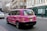A pink taxi with environmental advertising is driving on a city street. The vehicle has the words 'This cab is green' on its side, promoting renewable electricity from a company called Bulb. The street is lined with traditional European architecture buildings, and there are pedestrians walking by an urgent care center.