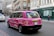 A pink taxi with environmental advertising is driving on a city street. The vehicle has the words 'This cab is green' on its side, promoting renewable electricity from a company called Bulb. The street is lined with traditional European architecture buildings, and there are pedestrians walking by an urgent care center.