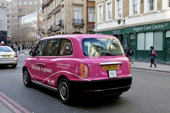 A pink taxi with environmental advertising is driving on a city street. The vehicle has the words 'This cab is green' on its side, promoting renewable electricity from a company called Bulb. The street is lined with traditional European architecture buildings, and there are pedestrians walking by an urgent care center.