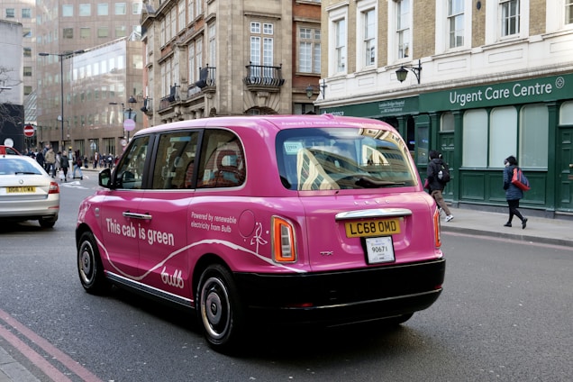 A pink taxi with environmental advertising is driving on a city street. The vehicle has the words 'This cab is green' on its side, promoting renewable electricity from a company called Bulb. The street is lined with traditional European architecture buildings, and there are pedestrians walking by an urgent care center.