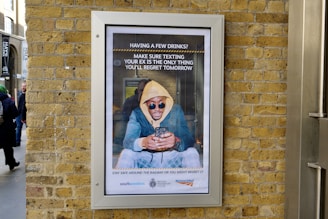 A framed advertisement on a brick wall shows a person in a yellow hoodie and sunglasses looking at their phone, accompanied by a message about being cautious when texting after drinking. The advertisement includes logos of transport organizations at the bottom, and people are passing by on the street.