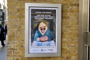 A framed advertisement on a brick wall shows a person in a yellow hoodie and sunglasses looking at their phone, accompanied by a message about being cautious when texting after drinking. The advertisement includes logos of transport organizations at the bottom, and people are passing by on the street.
