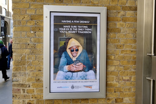 A framed advertisement on a brick wall shows a person in a yellow hoodie and sunglasses looking at their phone, accompanied by a message about being cautious when texting after drinking. The advertisement includes logos of transport organizations at the bottom, and people are passing by on the street.