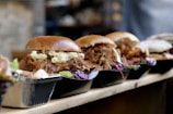 A row of delicious pulled pork sandwiches with macaroni and cheese, served in small black trays. The sandwiches have a glossy bun and are garnished with purple cabbage and lettuce.