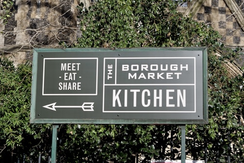 A green sign with white text that reads 'Meet - Eat - Share' and 'The Borough Market Kitchen'. An arrow points to the left, and the sign is surrounded by green foliage with a stone wall in the background.