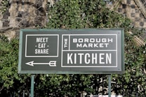 A green sign with white text that reads 'Meet - Eat - Share' and 'The Borough Market Kitchen'. An arrow points to the left, and the sign is surrounded by green foliage with a stone wall in the background.