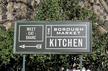 A green sign with white text that reads 'Meet - Eat - Share' and 'The Borough Market Kitchen'. An arrow points to the left, and the sign is surrounded by green foliage with a stone wall in the background.