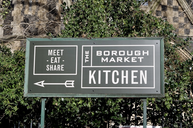 A green sign with white text that reads 'Meet - Eat - Share' and 'The Borough Market Kitchen'. An arrow points to the left, and the sign is surrounded by green foliage with a stone wall in the background.