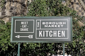 A green sign with white text that reads 'Meet - Eat - Share' and 'The Borough Market Kitchen'. An arrow points to the left, and the sign is surrounded by green foliage with a stone wall in the background.
