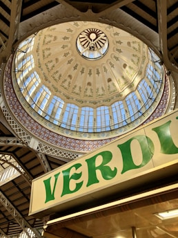 A beautifully ornate dome with intricate patterns and floral designs is adorned with decorative arches and a central circular skylight. There's a market stall sign with 'VERDU' written in bold green letters visible in the foreground. The structure displays an architectural mix of metal and glass.
