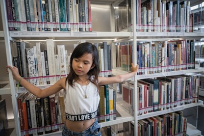 A young girl is standing in front of a bookshelf in a library. She has her arms extended to touch either side of the bookshelf. She is wearing a cropped white top with text on it and jeans. The bookshelves are filled with numerous books, organized and neatly arranged.