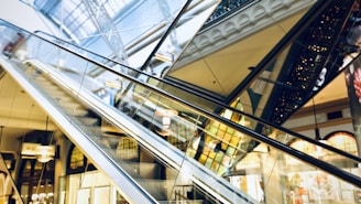 Several escalators crisscross each other inside a modern shopping mall. Large glass panels and an intricate ceiling structure are visible along with various building elements, including ornate railings and storefronts with lit displays.