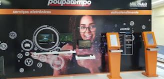 A wall display in a public service area with digital service sections. It features a large image of a smiling woman with glasses in the center, surrounded by digital screens and icons representing different electronic services. There are two orange kiosks with touch screens for self-service on the right side.