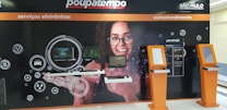 A wall display in a public service area with digital service sections. It features a large image of a smiling woman with glasses in the center, surrounded by digital screens and icons representing different electronic services. There are two orange kiosks with touch screens for self-service on the right side.