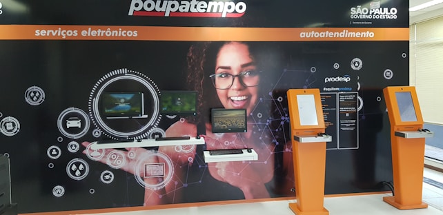 A wall display in a public service area with digital service sections. It features a large image of a smiling woman with glasses in the center, surrounded by digital screens and icons representing different electronic services. There are two orange kiosks with touch screens for self-service on the right side.