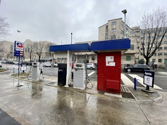 A gas station features multiple fuel pumps under a blue and red canopy. Vehicles are parked nearby, and the background shows urban buildings and leafless trees on an overcast day.