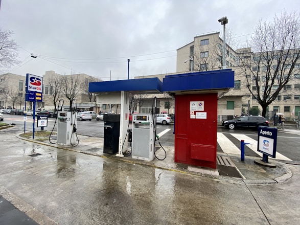 A gas station features multiple fuel pumps under a blue and red canopy. Vehicles are parked nearby, and the background shows urban buildings and leafless trees on an overcast day.