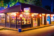 A vibrant street stall is illuminated with colorful lights and traditional lanterns under a dark blue evening sky. The stall has a wooden structure and is stocked with various goods, visible through large glass windows. Signs with Vietnamese text are displayed above the entrance.