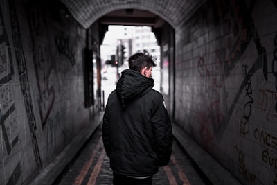 A model wearing a minimalist black jacket walking down a graffiti-lined alley.