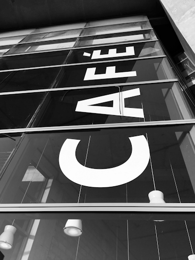 Large letters spelling out 'CAFÉ' displayed vertically on the glass facade of a modern building. The reflection of the structure and the sky is visible on the glass surface.