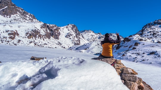person in blue jacket and orange pants sitting on rock formation covered with snow during daytime in Gosaikunda Nepal