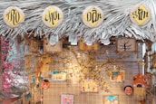A decorative stall with a thatched roof made of palm leaves, featuring circular signs with intricate black lettering on a yellow background. Various colorful art pieces and masks hang from the structure, surrounded by decorative flowers. The warm lighting and vibrant illustrations create a festive atmosphere.