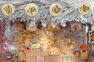 A decorative stall with a thatched roof made of palm leaves, featuring circular signs with intricate black lettering on a yellow background. Various colorful art pieces and masks hang from the structure, surrounded by decorative flowers. The warm lighting and vibrant illustrations create a festive atmosphere.