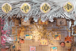 A decorative stall with a thatched roof made of palm leaves, featuring circular signs with intricate black lettering on a yellow background. Various colorful art pieces and masks hang from the structure, surrounded by decorative flowers. The warm lighting and vibrant illustrations create a festive atmosphere.