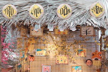 A decorative stall with a thatched roof made of palm leaves, featuring circular signs with intricate black lettering on a yellow background. Various colorful art pieces and masks hang from the structure, surrounded by decorative flowers. The warm lighting and vibrant illustrations create a festive atmosphere.