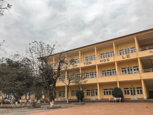 A three-story school building with a yellow facade and multiple uniform windows. The central structure has large letters on the facade, and there are some leafless trees in the foreground with benches positioned nearby. The sky is overcast, conveying a late autumn or winter atmosphere.
