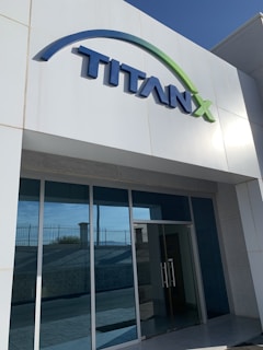 A modern building entrance with a large sign that reads 'TITANX' in bold, blue letters. The building has a sleek, minimalist design with glass doors and a glass facade. The sign features a green arc above the name.