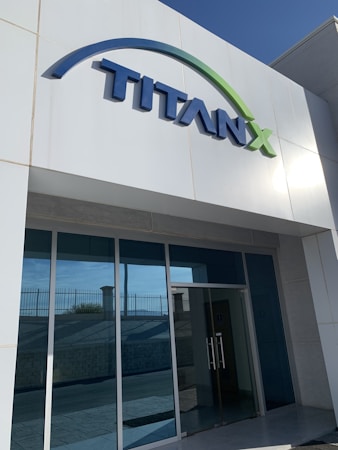 A modern building entrance with a large sign that reads 'TITANX' in bold, blue letters. The building has a sleek, minimalist design with glass doors and a glass facade. The sign features a green arc above the name.