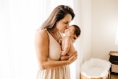 A woman gently holds a newborn baby close to her chest, tenderly kissing the baby's forehead. The setting is a softly lit room with sheer curtains allowing natural light to illuminate the scene. A wicker bassinet is visible in the background.