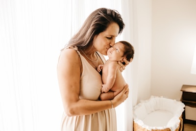 A woman gently holds a newborn baby close to her chest, tenderly kissing the baby's forehead. The setting is a softly lit room with sheer curtains allowing natural light to illuminate the scene. A wicker bassinet is visible in the background.