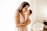 A woman gently holds a newborn baby close to her chest, tenderly kissing the baby's forehead. The setting is a softly lit room with sheer curtains allowing natural light to illuminate the scene. A wicker bassinet is visible in the background.