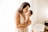 A woman gently holds a newborn baby close to her chest, tenderly kissing the baby's forehead. The setting is a softly lit room with sheer curtains allowing natural light to illuminate the scene. A wicker bassinet is visible in the background.