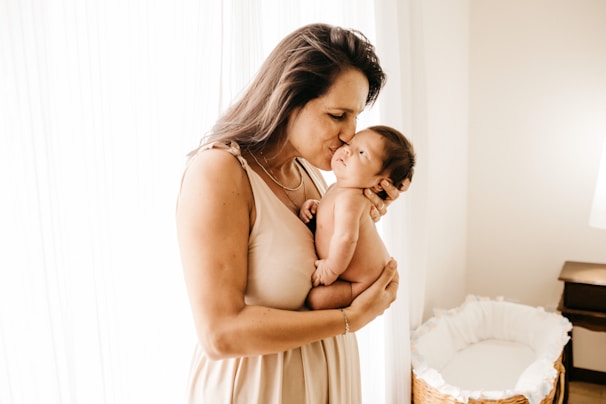 A woman gently holds a newborn baby close to her chest, tenderly kissing the baby's forehead. The setting is a softly lit room with sheer curtains allowing natural light to illuminate the scene. A wicker bassinet is visible in the background.