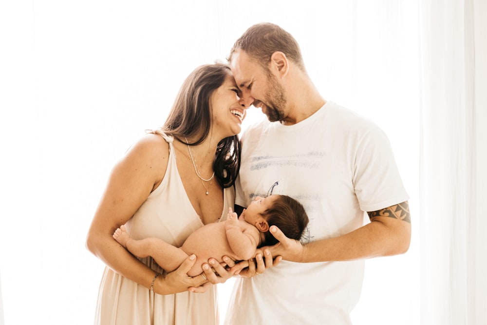 man in white crew neck t-shirt kissing woman in white dress with a baby on their hands