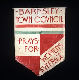 A fabric banner with bold red text on a white background. The text reads 'Barnsley Town Council Prays for Women's Suffrage'. The design features a combination of horizontal and diagonal sections with a prominent red and light green stripe.