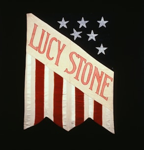 A vertically striped flag with red and white stripes, featuring the name 'Lucy Stone' in bold red letters on a diagonal white banner against a black background adorned with silver stars.