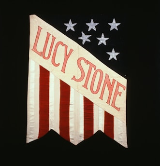 A vertically striped flag with red and white stripes, featuring the name 'Lucy Stone' in bold red letters on a diagonal white banner against a black background adorned with silver stars.