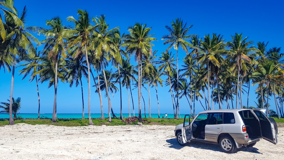 A bright turquoise Toyota Yaris parked by a sunlit Curacao beach with palm trees swaying.