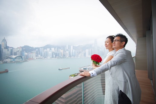 A couple dressed in wedding attire stands on a balcony overlooking a large body of water and a city skyline. The bride holds a bouquet of red roses, and both appear content, gazing into the distance with a serene expression. The sky is overcast, with a mix of clouds, and the architecture of the distant skyscrapers suggests a modern urban environment.